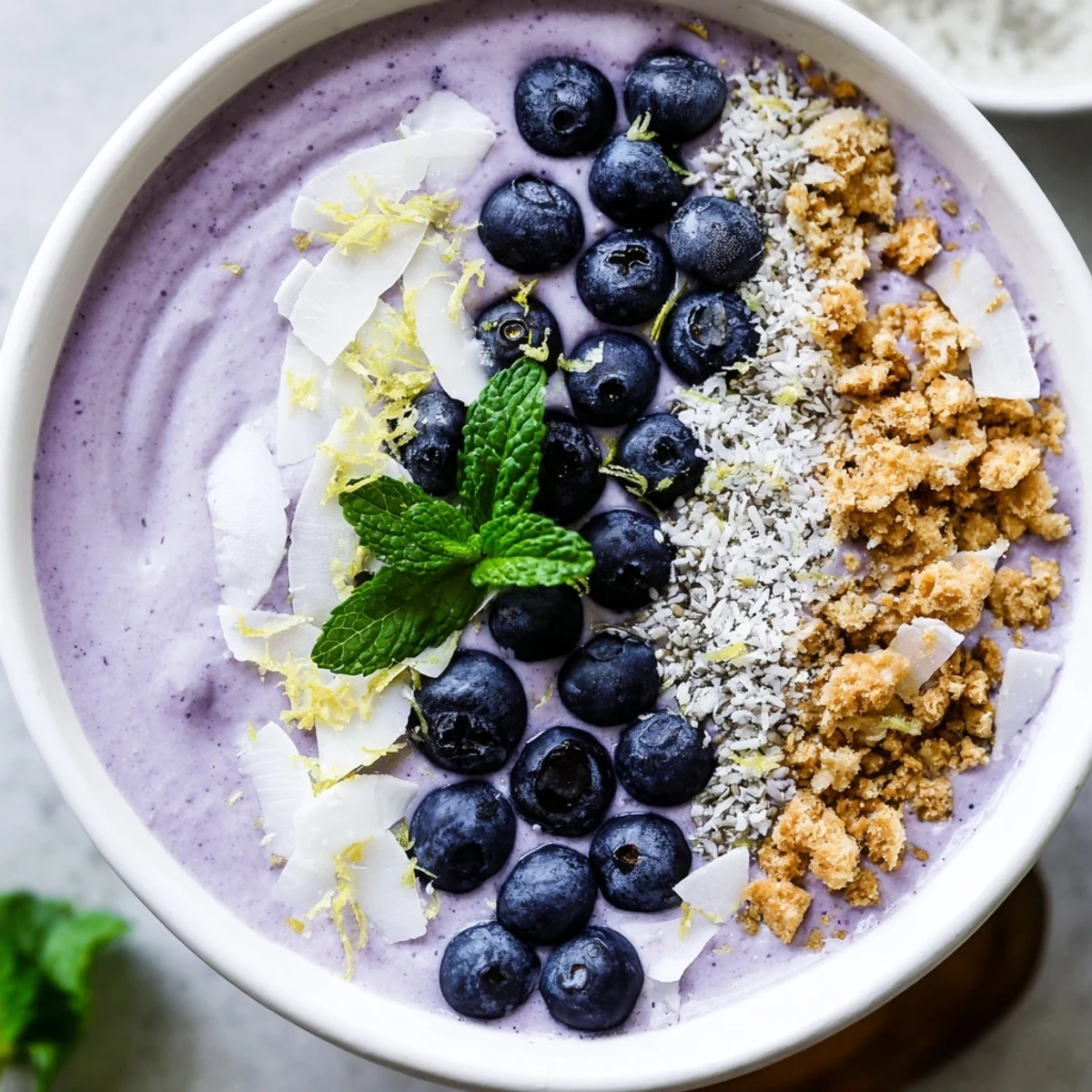 Bright and creamy Lemon Blueberry Smoothie Bowl in a white bowl, garnished with lemon zest and granola clusters.