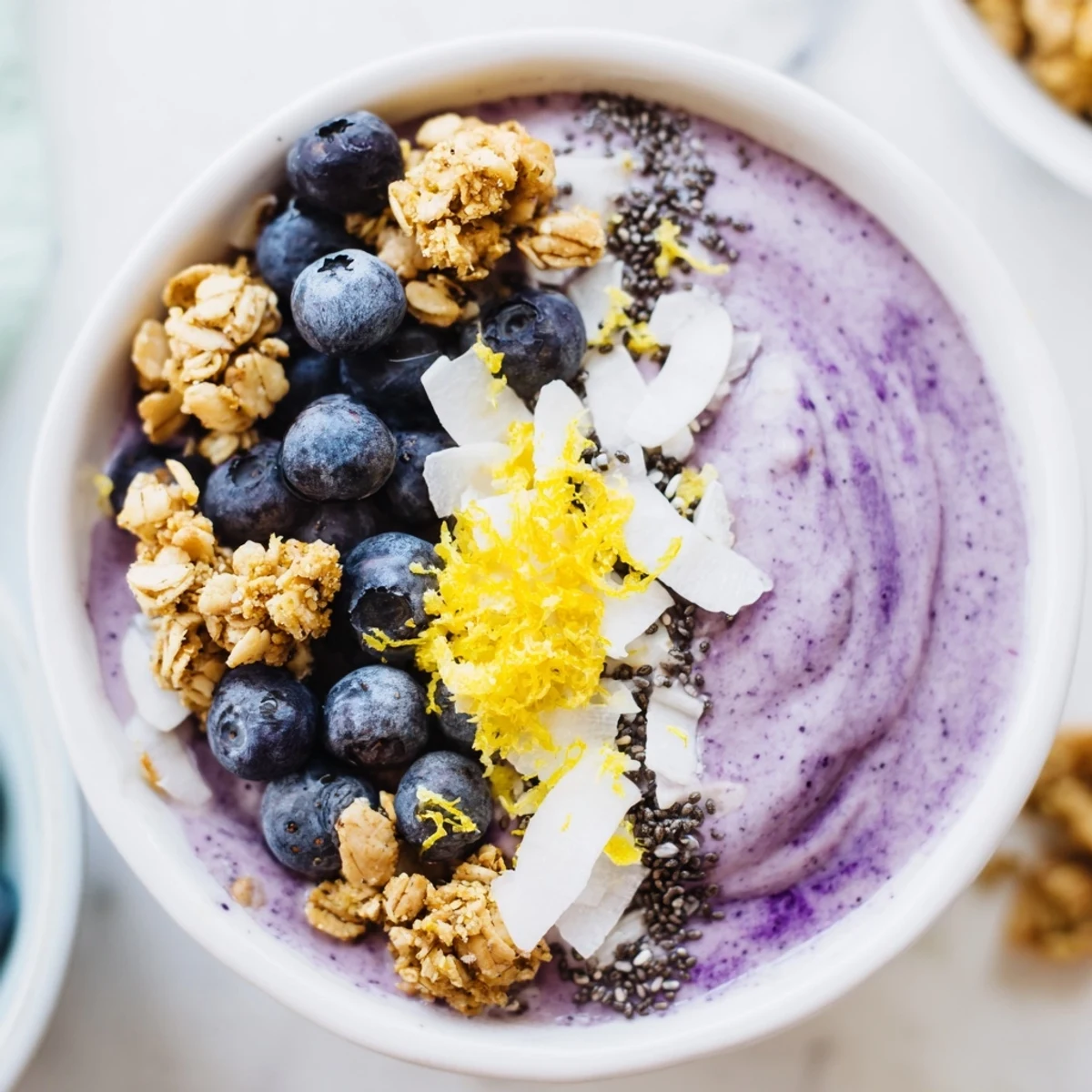 Spoon-ready Lemon Blueberry Smoothie Bowl topped with crunchy granola and coconut flakes on a marble counter.