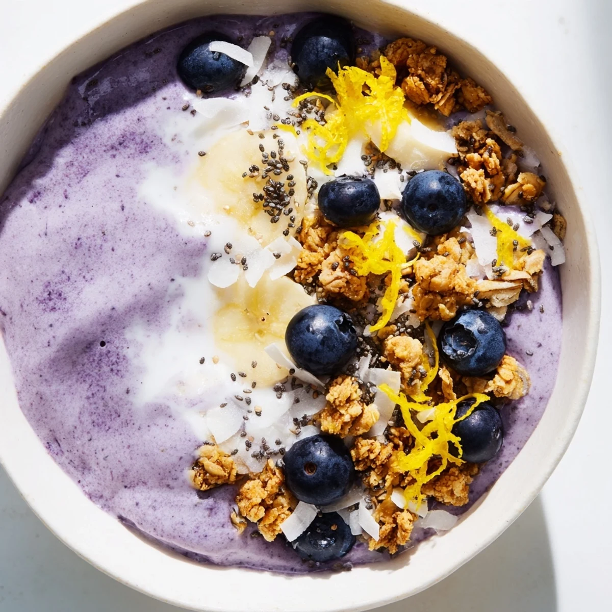 Close-up of a creamy Lemon Blueberry Smoothie Bowl with lemon zest garnish beside a glass of green tea.