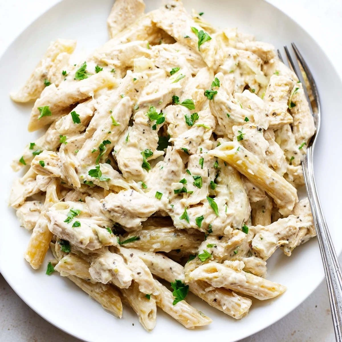 A close-up photo of Crockpot Garlic Parmesan Chicken Pasta with shredded chicken and creamy sauce coating penne, garnished with fresh parsley.