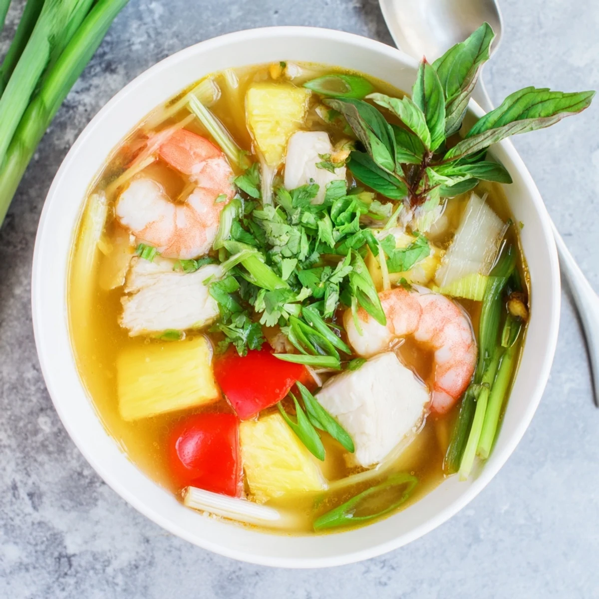A close-up of Canh Chua soup in a white bowl, showcasing tender fish chunks and pink shrimp swimming in a vibrant, orange-hued broth. The glossy surface of the soup reflects the light, with visible slices of pineapple, tomato wedges, and fresh cilantro leaves floating on top. Steam gently rises, suggesting it's freshly cooked. The background is a simple, light-colored marble countertop, keeping the focus entirely on the steaming bowl of Vietnamese sweet and sour soup.