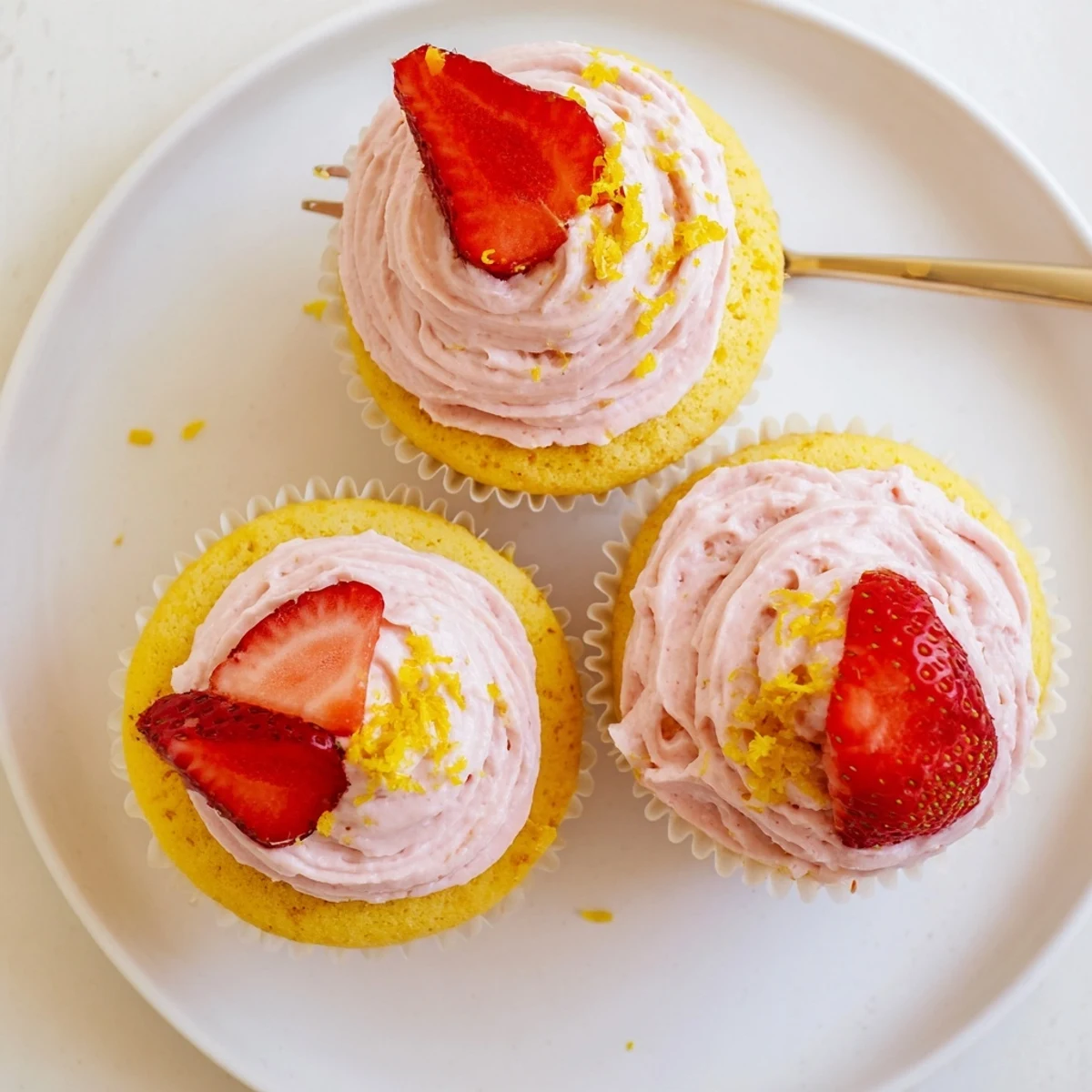 Strawberry lemonade cupcakes topped with pink strawberry frosting and fresh strawberry halves on a white plate