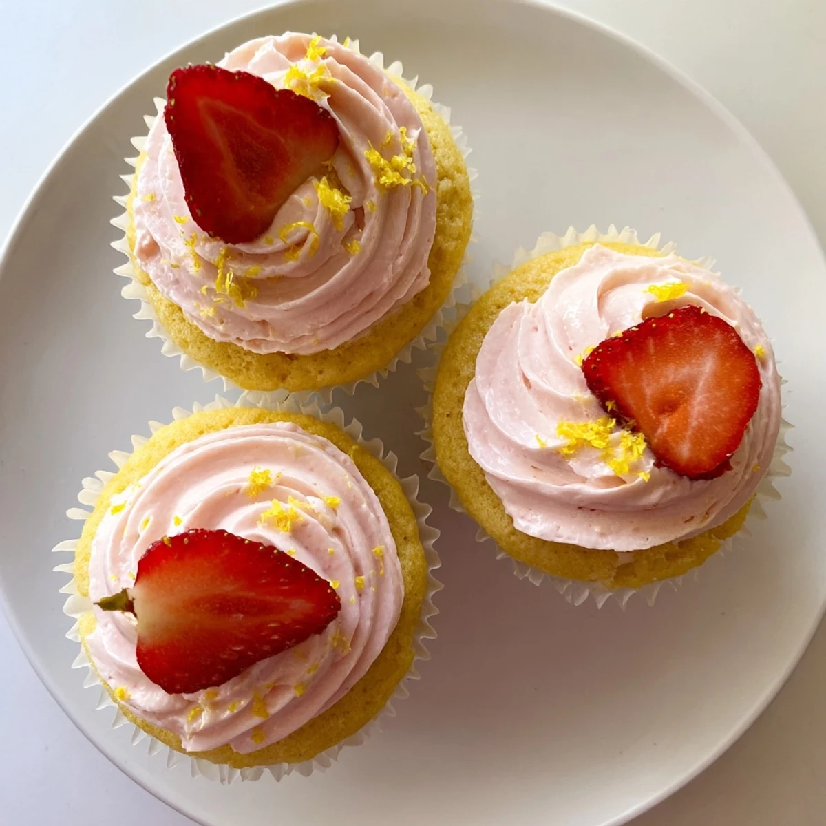 Golden lemon cupcakes piped with creamy pink strawberry frosting and garnished with lemon zest and berries